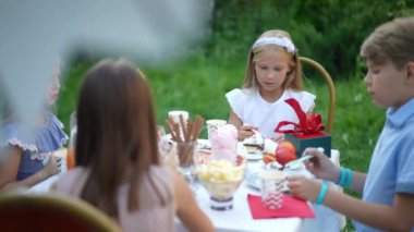 Beyaz elbiseli sarışın kız arka bahçede arkadaşlarıyla oturup doğum gününü kutluyor. Rahatlamış, mutlu beyaz bir çocuğun portresi. Açık havada eğleniyor ve lezzetli yemekler yiyor.
