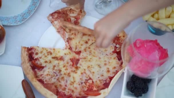屋外のテーブルの上でスローモーションでピザスライスを区切るトップビューの子供の手 近くで認識できない白人の女の子は 裏庭のピクニックでパーティーを祝うプレートからおいしい食べ物を取って — ストック動画