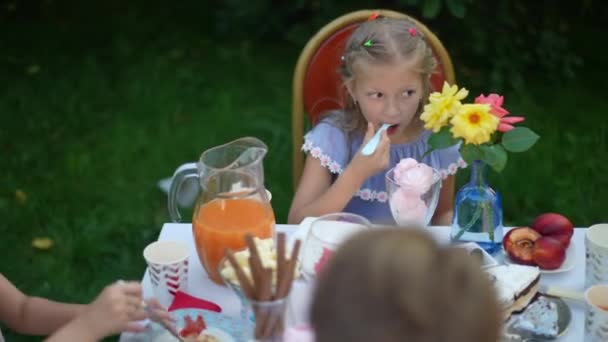 青いドレスの白人の女の子は 屋外のテーブルで友人と座っておいしい誕生日ケーキを楽しんでいます 高角度ビューポートレートのブロンド子供とともに茶色の目は裏庭のパーティーでおいしい甘い食べ物を食べる — ストック動画