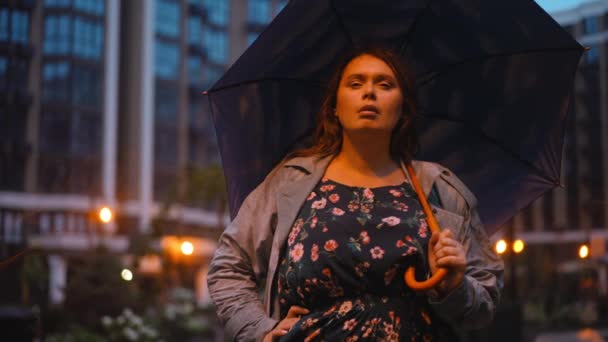 遠くを見て 夜の街路灯の中に立ってカメラを見て傘を持つ白人プラスサイズの女性の肖像画 自信を持って魅力的な大人の女性ポーズで雨の都市部屋外 — ストック動画