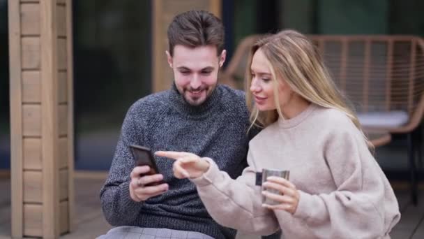 スマートフォンアプリでソーシャルメディアをスクロールして話している楽しい白人の大人のカップル笑顔 正幸せなリラックスした白人のボーイフレンドとガールフレンド楽しみます休暇チャット屋外で座って — ストック動画
