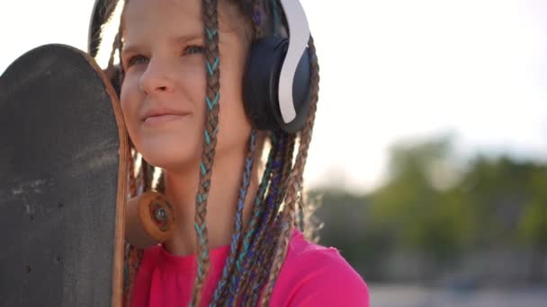一个头像是戴着耳机 自信而又积极的少女 她在户外站在阳光下滑板 开心的笑着的白人青少年 在慢动作中欣赏着早晨的音乐 — 图库视频影像