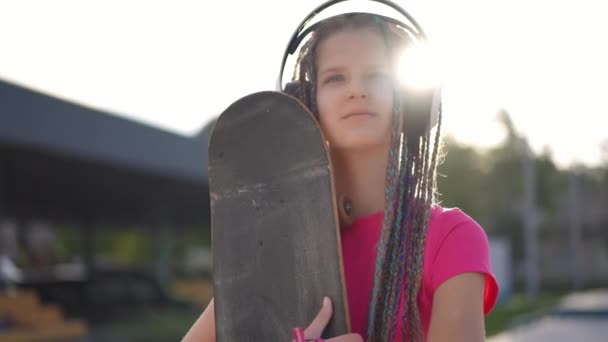 ヘッドフォンの魅力的な自信のある十代の女の子の肖像は笑顔を離れて見てスケートボードを抱きしめる ポジティブな白人ティーンスタンディングで日の出楽しい趣味 — ストック動画