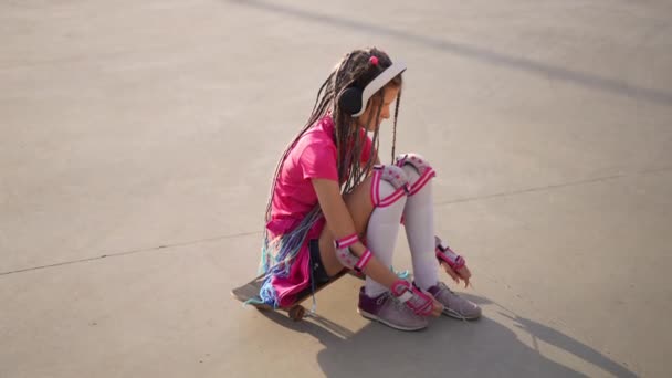 高角度广角镜头的白人少女与辫子系鞋带慢动作坐在滑板上 自信的青少年在耳机里听着音乐准备滑板 — 图库视频影像