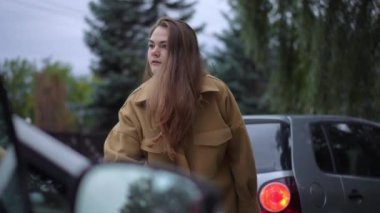 Kızgın sinirli genç kadın dışarıda hasarlı bir arabaya bakarken başka tarafa bakıyor. Bulutlu bir günde trafik kazasında tatminsiz beyaz kadın portresi