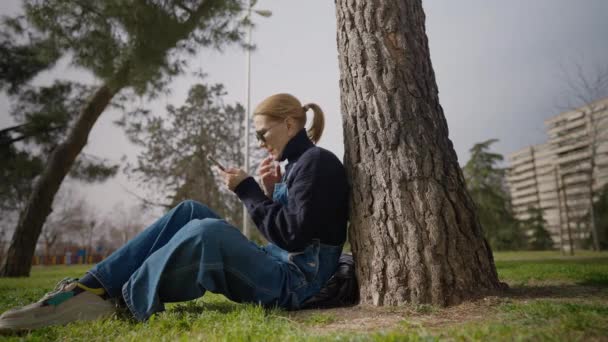 戴着太阳镜的多愁善感的女人坐在绿茵的草地上靠在树上 用智能手机应用在网上发短信 白种人自信的女士在户外享受周末休闲的开阔视野 — 图库视频影像