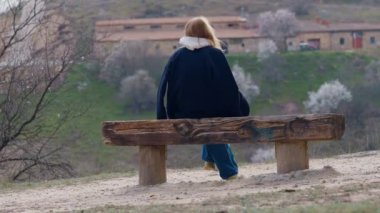 Soğuk bahar gününde bankta oturup düşünen geniş açılı bir bakış açısı olan kadın. Tanımlanamayan sarışın beyaz kadın hafta sonu açık havada eğleniyor.