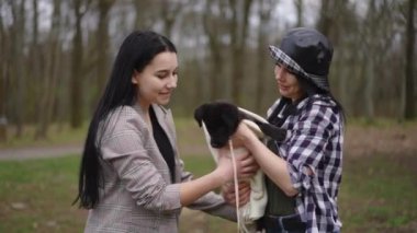 Gülümseyen beyaz kadının orta boy portresi köpek yavrusunu arkadaşına uzatırken çekilmiş. Mutlu, rahat, kendine güvenen arkadaşlar hafta sonu evcil hayvanla parkta eğleniyor.