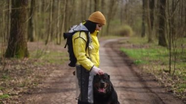 Geniş açı, mutlu, kendine güveni tam, park alanında oturan büyük köpeğin kafasını okşayan bir kadın. Açık havada yağmurluk giymiş, gülümseyen beyaz kadın.