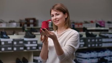 Orta boylu, gülümseyen beyaz bir kadın kırmızı yüksek topuklu sandaleti inceliyor. Ayakkabı mağazasında yeni ayakkabılar seçen düşünceli, kendine güvenen bir kadının portresi.