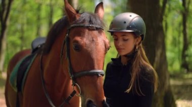 Genç ve çekici bir kadın güneşli ormanda hayvanla ayakta duran zarif bir atın yüzünü ve yelesini okşuyor. Dışarıda güneş ışığıyla beslenen kasklı, kendine güveni tam beyaz atlı bir at. Yavaş çekim