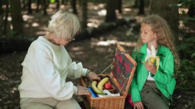 Rahat yaşlı kadın ve genç kız ormanda piknikte oturan meyveleri seçiyorlar. Kafkasyalı mutlu büyükanne ve torunun orta boy portresi. Muz ve elma vitamini yiyorlar.