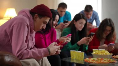Bulanık odaklı arkadaşlarıyla telefon uygulamalarında internette sörf yapan konsantre genç bir adamın yan görünümü. Genç Kafkasyalı erkek ve kadınlar sosyal medyada kayıyor, içeride parti yapıyorlar.