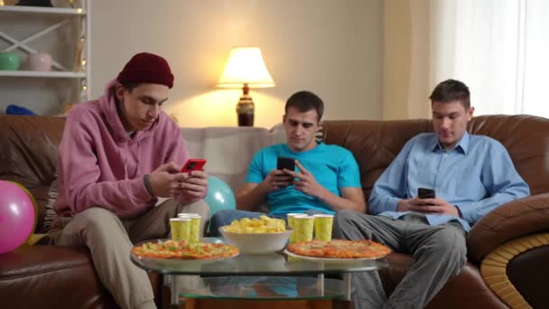 3人のリラックスした吸収された若い白人男性は ソーシャルメディアアプリケーションをスクロールして テーブルの上のジャンクフードでソファーに座っている 週末のレジャーミーティングを楽しんでいるケアフリーの男性の友達 — ストック動画