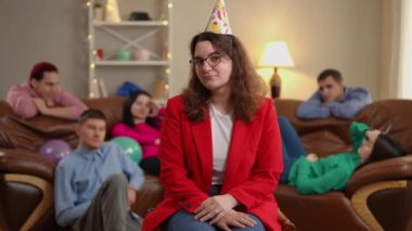 Parti şapkalı ve gözlüklü genç bir kadın arka planda sıkılmış arkadaşlarıyla iç çekiyor. Evdeki sıkıcı partide tatmin olmamış Kafkas doğum günü kadınının portresi.