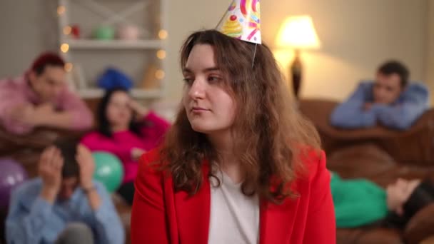 一个头戴派对帽的无聊女人的画像 她坐在室内 后面是疲惫的朋友 在室内度过无聊假期的白人加身材女士 — 图库视频影像