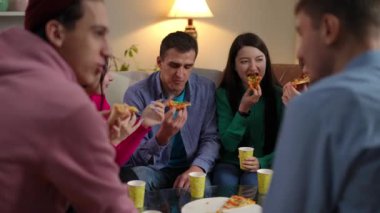 Bir grup kaygısız genç arkadaş yavaş çekimde konuşuyor oturma odasında oturmuş pizza yiyorlar. Kafkasyalı kadın ve erkekler içeride eğlenmekten zevk alıyor.