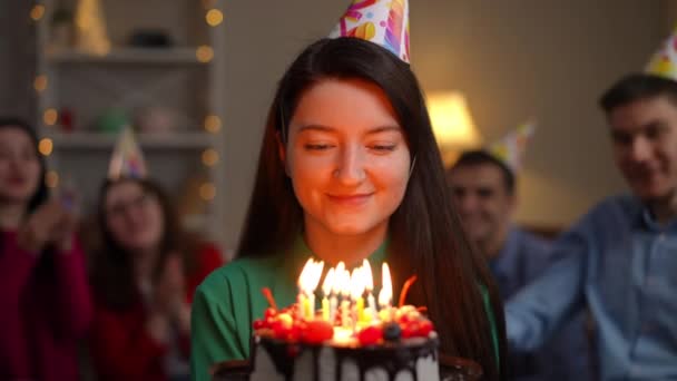 年轻迷人的女人戴着派对帽 在生日蛋糕上吹灭蜡烛 看着相机 模糊的朋友圈在背后欢欣鼓舞 快乐的女士在室内摆设甜点 — 图库视频影像