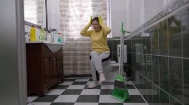 Migreni olan yorgun bir kadın tuvalette oturmuş ellerini tutarak başını tutuyor. Kafkasyalı genç ev kadınının ev içi baş ağrısı çeken portresi.