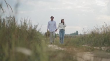 Geniş açılı genç adam, eşini ceketle saklayan bir kadınla dolaşıyor ve partneriyle birlikte ayrılıyor. Mutlu, kendine güvenen beyaz bir çift güz tarlasında yürüyor.