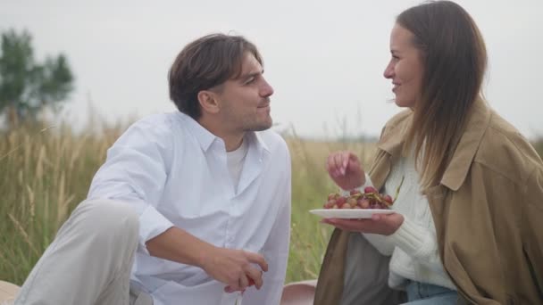 微笑迷人的女人用葡萄喂男人 慢吞吞地吃水果 白种人年轻夫妇在阴天的户外秋田里享受野餐的快乐无忧无虑的画像 — 图库视频影像