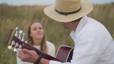 Yan görünüm: Piknikte arka planda oturan bulanık bir kadınla gitar çalan yetenekli genç adam. Bulutlu bir günde buluşup eğlenen mutlu beyaz çift.