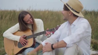 Güz tarlasında piknikte şarkı söyleyen bulanık bir adam gibi gitar çalan gülümseyen yetenekli genç bir kadının portresi. Olumlu, rahat, Kafkasyalı çift boş zamanların ve hobilerin tadını çıkarıyor.