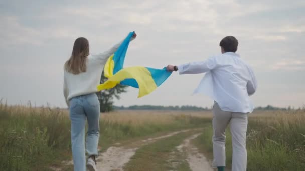 ウクライナ国旗を伸ばすフィールドでゆっくりと動く楽しいカップルの追跡ショット ワイドショットバックビュー幸せな陽気な若者と女性が屋外で喜ぶ — ストック動画
