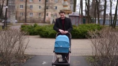 Geniş açıda gülümseyen adam, park yolunda bebek arabasıyla yürüyen bebeğe hayran. Dolly, bulutlu bir sabahta gezen mutlu beyaz bir babanın portresi.