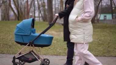 Park sokağında mavi bebek arabasıyla yürüyen kendine güvenen bir çiftin yan görüntüsü. Kafkasyalı erkek ve kadın bulutlu bir sabahta yeni doğmuş çocukla geziyorlar. Huzurun tadını çıkarıyorlar.