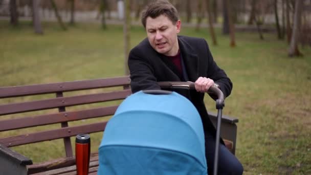 不満を抱いた男性は 公園のベンチに座っているベビーカーを揺さぶる新生児と話している 落ち着く子供を試みている手で耳を覆う疲れた白人父の肖像画 — ストック動画