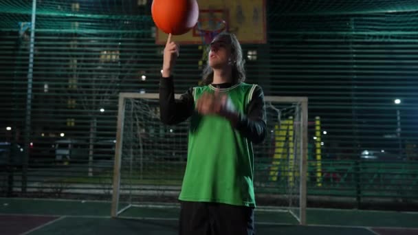ブルネットの若い男が夜にスポーツグラウンドに立って指にバスケットボールボールを回転する中型ショット 集中した白人スポーツマンの肖像画 屋外で遊ぶトレーニング — ストック動画