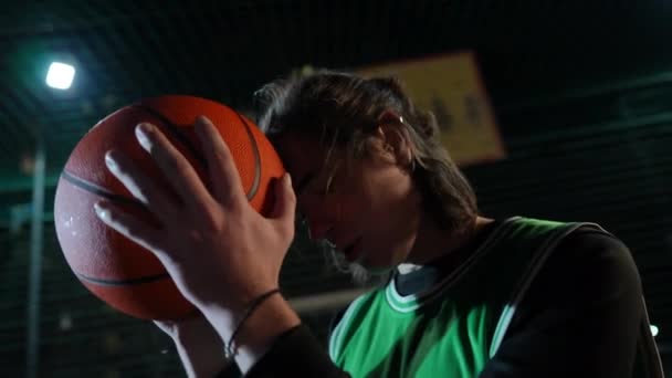 疲れた若いスポーツマンは 手でバスケットボールのボールに大きく頭を傾けています 疲れ果てたコーカサス人の肖像画 屋外の夜のスポーツ用品 — ストック動画