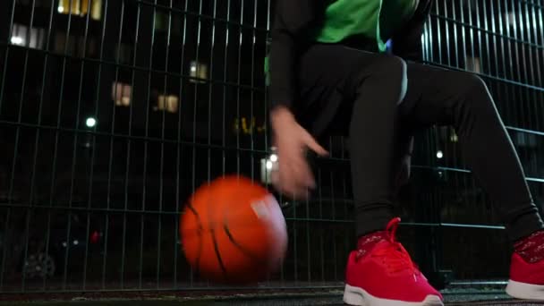 无法辨认的运动员的腿跳起球 靠在网状栅栏前 然后走开了 年轻健康的白人男子在夜间城市的运动场上打篮球 带着装备离开 — 图库视频影像