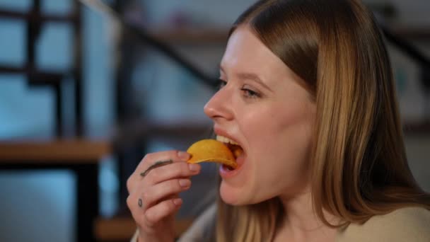 屋内でポテトチップスを食べている若い女性のクローズアップ顔 自宅でファーストフードを楽しんでいるコーカサス女性のヘッドショット側の角度のビューの肖像画 — ストック動画