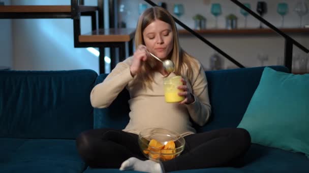 若い妊娠した白人女性は 甘い蜂蜜と塩辛いチップを食べて カメラを見つめている 自宅のリビングルームでソファーにポーズする魅力的な期待の混合食品のフロントビューの肖像画 — ストック動画