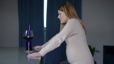 Yan bakış depresif hamile kadın mutfakta dikilip bir bardak kırmızı şaraba bakıyor. Evde bağımlılıkla savaşan üzgün Kafkasyalı beklentisi.