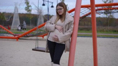 Düşünceli, üzgün, hamile bir kadın sütununa yaslanmış, çocuk parkına bakıyor. Açık havada bulutlu bir sonbahar gününde genç beyaz beklenen düşüncelerin portresi.