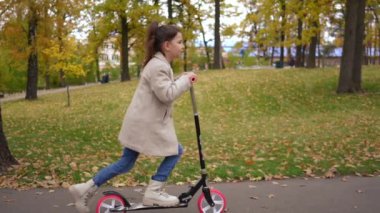 Yavaş çekimde gülümseyerek scooter 'a binen güzel bir kızın yan görüntüsü. Kendine güveni tam, beyaz bir çocuk sonbahar parkında eğleniyor.