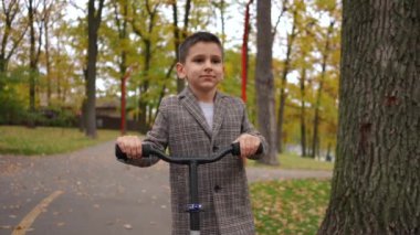 Ağır çekimde gülümseyen ve scooterla yürüyen beyaz bir çocuğun Dolly portresi. Ön manzara kendinden emin beyaz bir çocuk sonbahar günü açık havada park alanında geziniyor.
