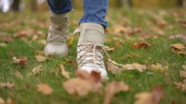 Ön manzara, beyaz çizmeli kadın çocuk ayakları yeşil çimlerin üzerinde, düşmüş sarı ve kırmızı yapraklarla yürüyor. Dolly, sonbahar parkında gezinirken, hafta sonu tatilinin keyfini çıkarırken, tanınmaz halde yakın çekim yaptı.