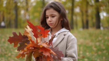Düşmüş yapraklardan oluşan buketi olan sevimli bir kız yavaşça el sallayarak uzaklaşıyor. Sonbahar parkında kendine güveni tam bir beyaz çocuk.