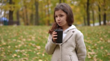 Yavaş çekimde kağıt bardaktan çay içen çekici bir kızın portresi çayıra giren bir çocukla içkiyi paylaşıyor. Beyazlı olumlu kız ve erkek kardeşler, sonbahar haftasonu dışarıda.