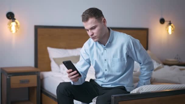 Poważny Młody Człowiek Siedzi Łóżku Przewijając Ekran Dotykowy Smartfona Zwolnionym — Wideo stockowe