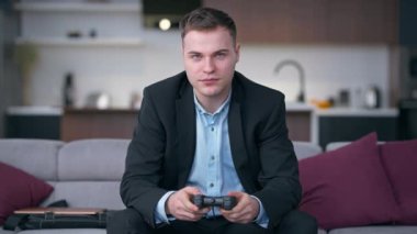Ön görüş takım elbiseli genç adam joystickle oynuyor yavaş çekimde başarıyı işaret ediyor kanepeye yaslanıyor. Kafkasya 'nın mutlu başlangıcının orta boy portresi evde keyif çatıyor.