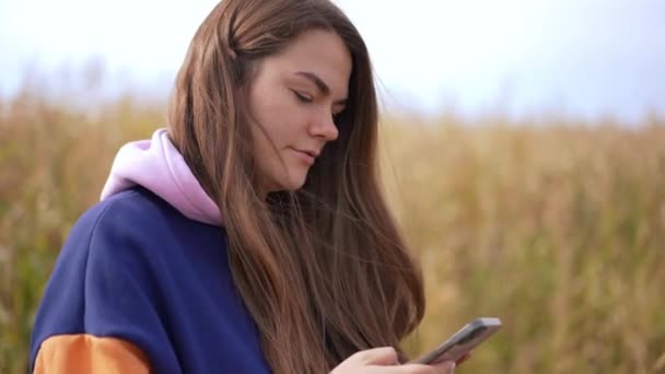スマートフォンでソーシャルメディアアプリをスクロールする若い女性のサイドビューの肖像画 アウトドアで秋のフィールドでカカシアの魅力的な自信を持った女性 — ストック動画