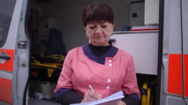 Kadın bir sağlık görevlisi ambulansın arka planında duruyor ve hastaların tıbbi geçmişini kağıda yazıyor.