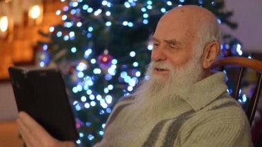 Yaşlı bir adam, güzel, ışıl ışıl bir Noel ağacının önünde bir video görüşmesinde konuşuyor.