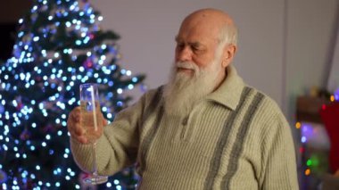 Yaşlı, kır saçlı, sakallı bir adam elinde bir bardak beyaz şarapla bir Noel ağacının yanında duruyor.