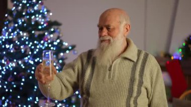 Bir bardak beyaz köpüklü şarapla yaşlı, kır saçlı bir adam Noel ağacının yanında Noel için kadeh kaldırır.